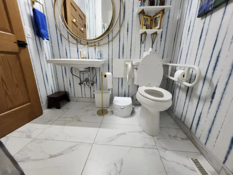 Photograph of a bathroom remodel by Adaptive Remodeling Solutions, showing a white toilet with adjustable grab bars next to a wall mounted sink and gold mirror.