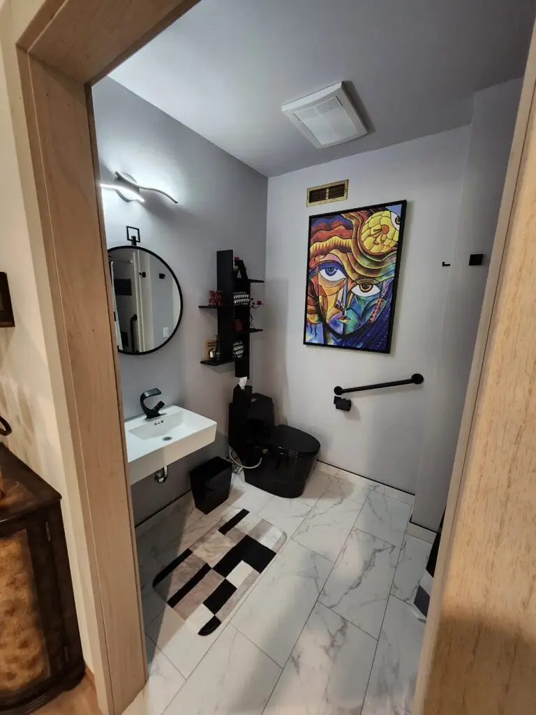 Photograph of a modern bathroom remodel completed by Adaptive Remodeling Solutions, featuring black accents, black toilet, black grab bar, mirror, and white wall mounted sink with a black handle.