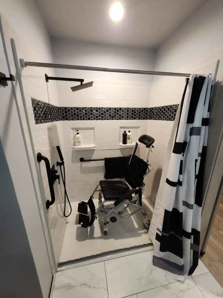 Photograph of a modern zero entry shower remodel completed by Adaptive Remodeling Solutions complete with detachable nozzle, large flat shower head, and grab bars. Inside the shower is a wheelchair.