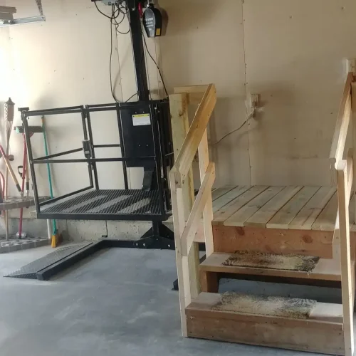 Photograph of a wheelchair lift next to a set of wooden stairs inside a garage, installed by Adaptive Remodeling Solutions.